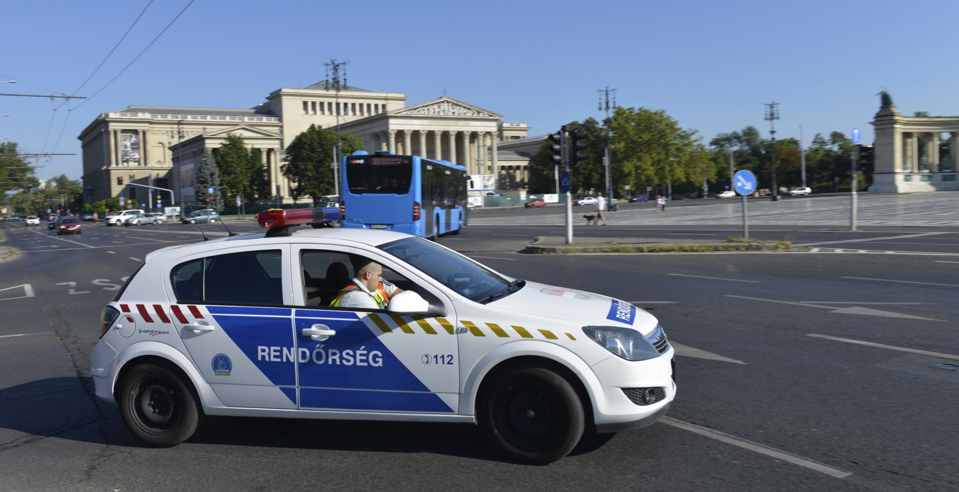 Magyar rendőrautó halad a budapesti Hősök tere mellett, szimbolizálva a közúti biztonságot és az Európai Unió által előírt új, kötelező autó felszerelések ellenőrzését a magyar közutakon.