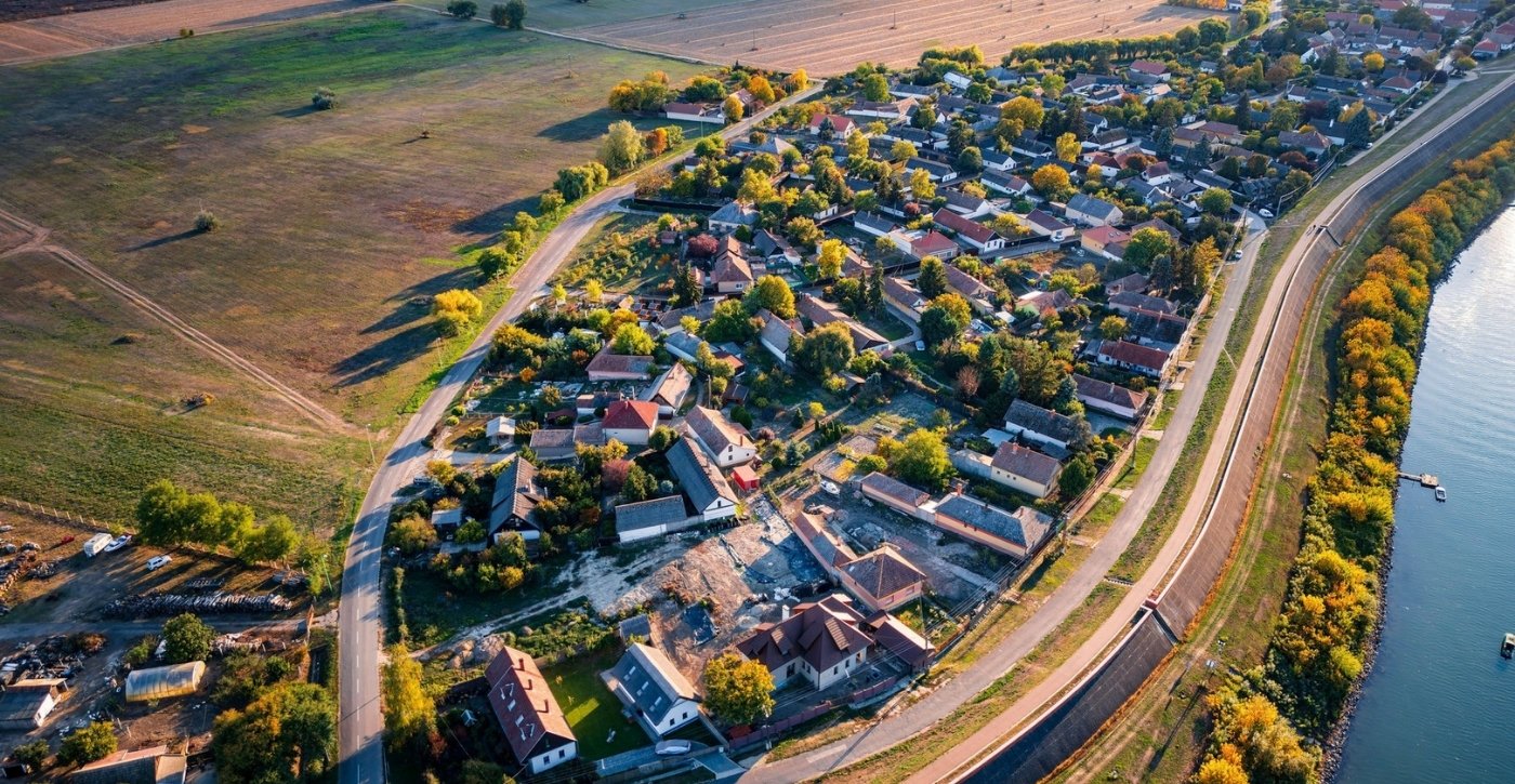 Légi felvétel egy festői magyar faluról, amelyet a kanyargós Duna határol. A falu házai sűrűn helyezkednek el a folyó mentén, zöld mezőkkel és erdőkkel körülvéve.