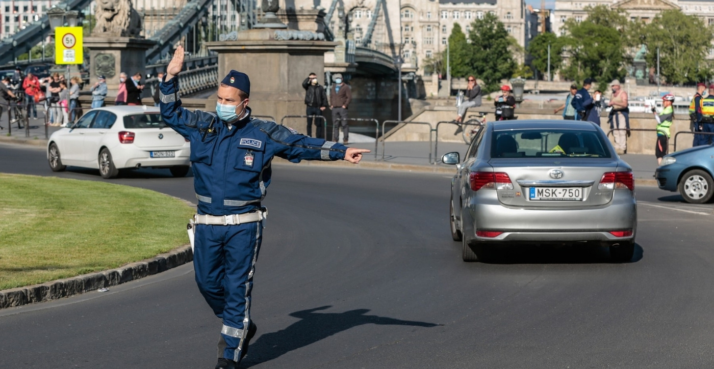 Egy rendőregyenruhát és orvosi maszkot viselő rendőr karjelzésekkel irányítja a forgalmat egy forgalmas budapesti csomópontban, a háttérben a Széchenyi lánchíd látható; a kép a KRESZ-szabályok módosítását és a közúti bírságok szigorítását szemlélteti.