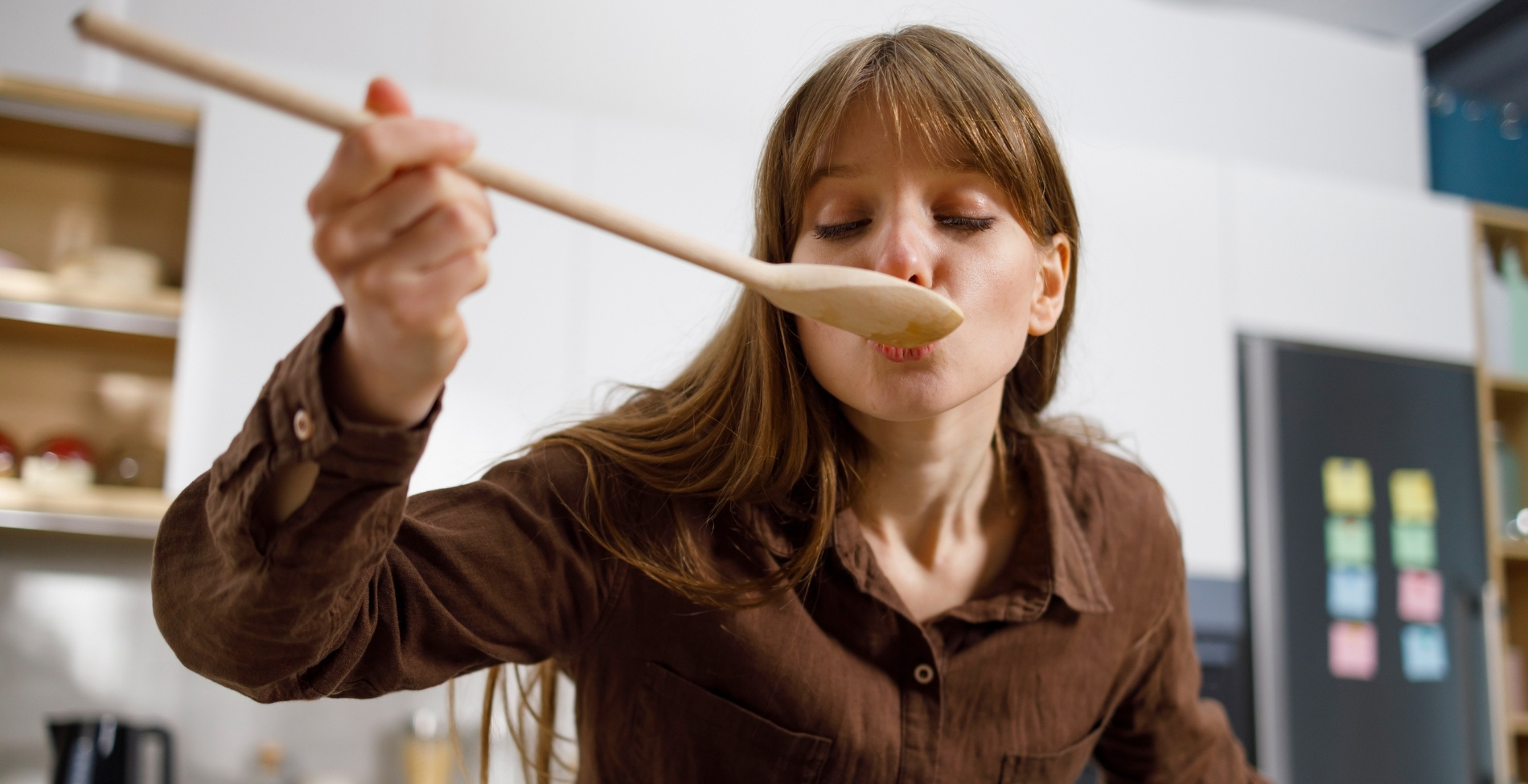 Fiatal nő csukott szemmel, élvezettel kóstol meg egy ételt egy fakanálból a konyhában, miközben azon gondolkodik, mi legyen a mai magyar étel, vagy éppen a tökéletes lángos titkát kutatja a „mit egyek ma” kérdésre válaszolva.