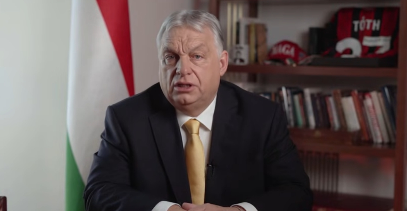 Orbán Viktor miniszterelnök elszánt arckifejezéssel, ünnepi öltözetben beszél a magyar zászló mellett, miközben a háttérben látható személyes tárgyak és könyvespolc egy közvetlen, mégis fajsúlyos politikai üzenet hangulatát árasztják.